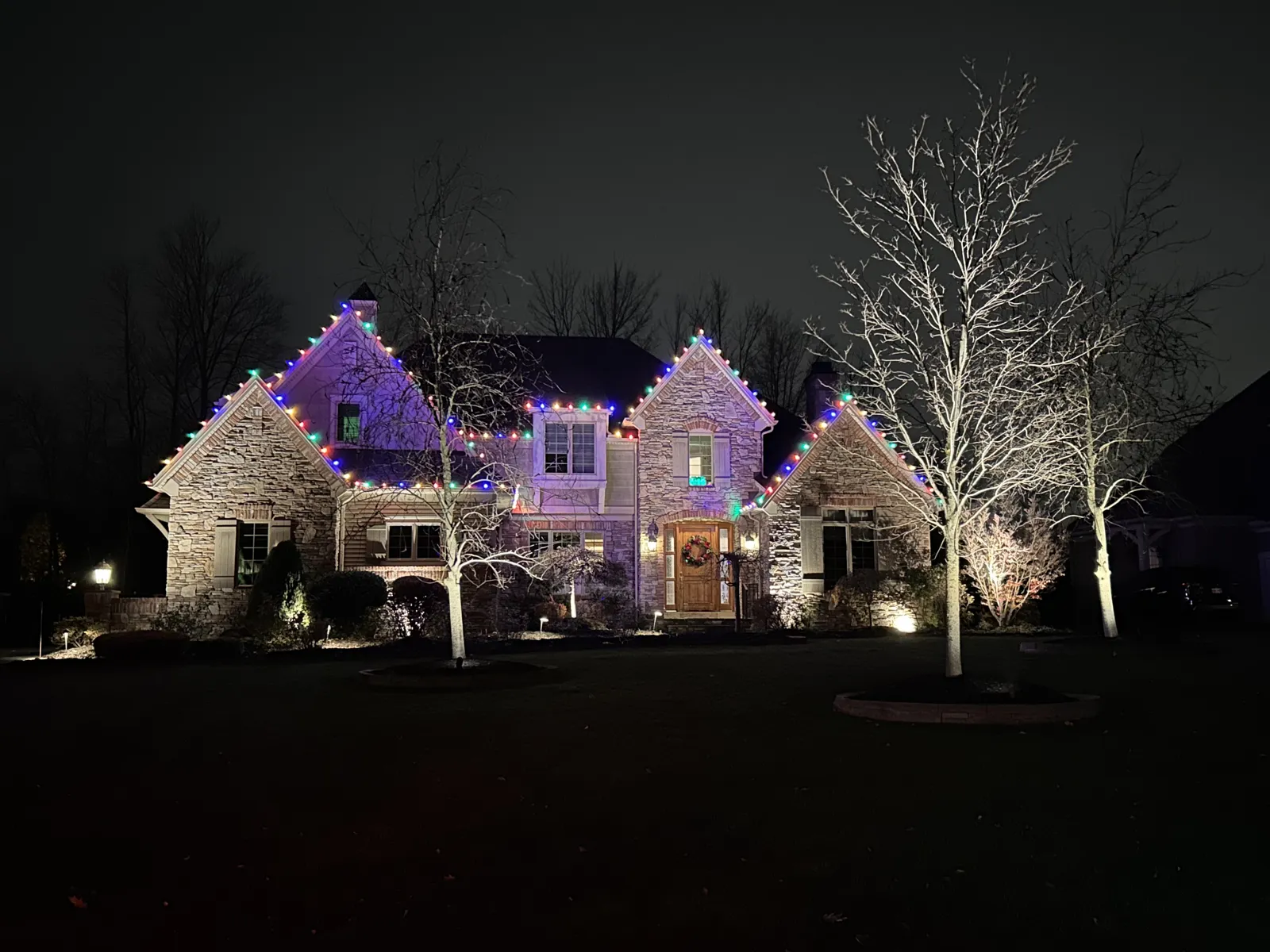 Stone home with multi-color roofline lights and warm-white wrapped trees