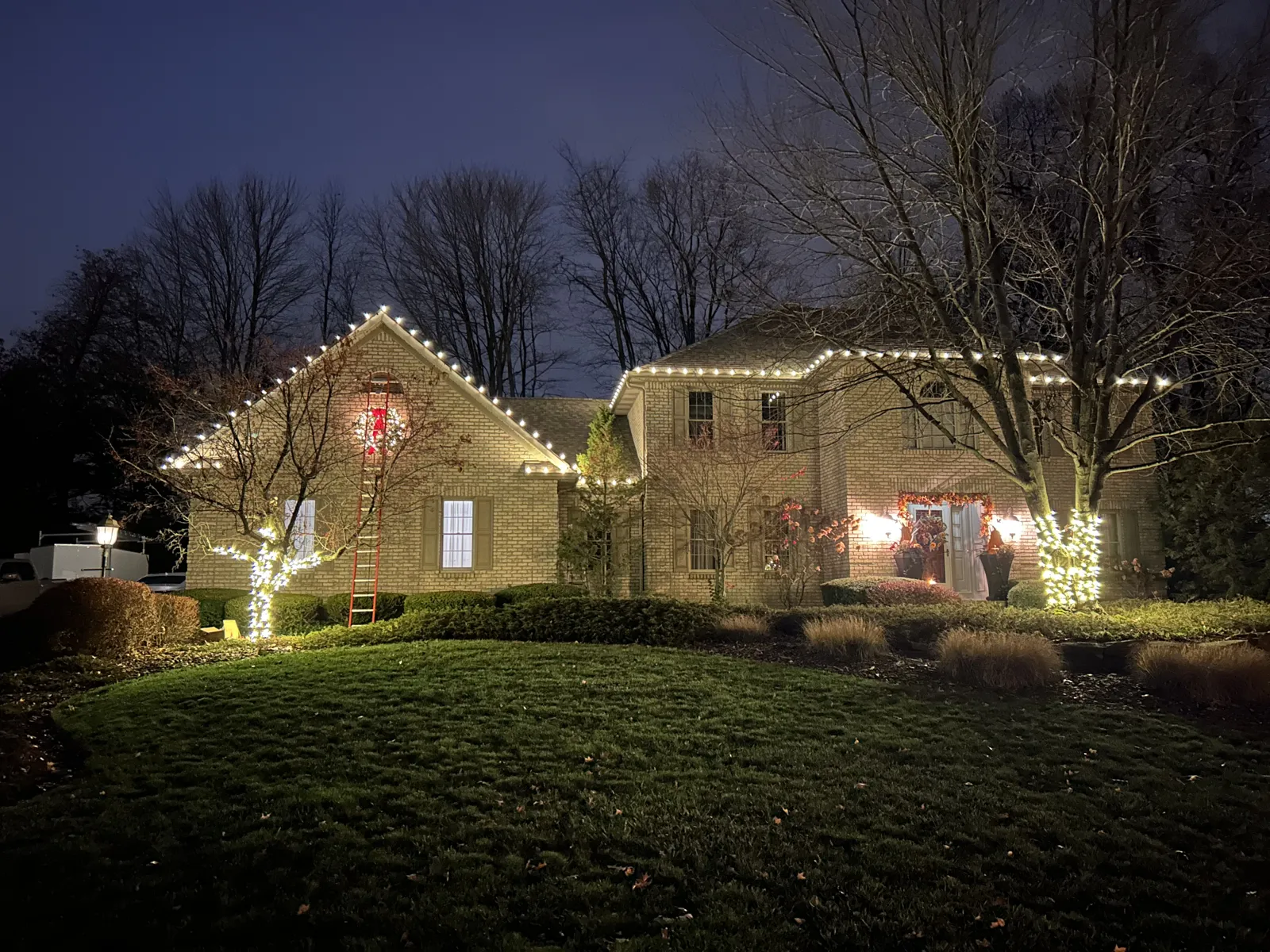 Two-story home with warm-white roofline lights and wrapped trees at dusk