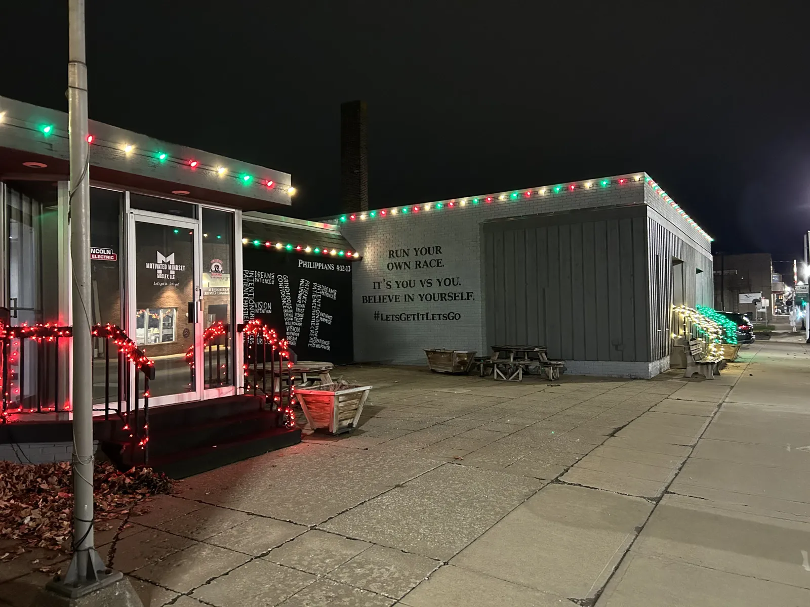 Storefront with red and green Christmas lights beside a BELIEVE mural at night